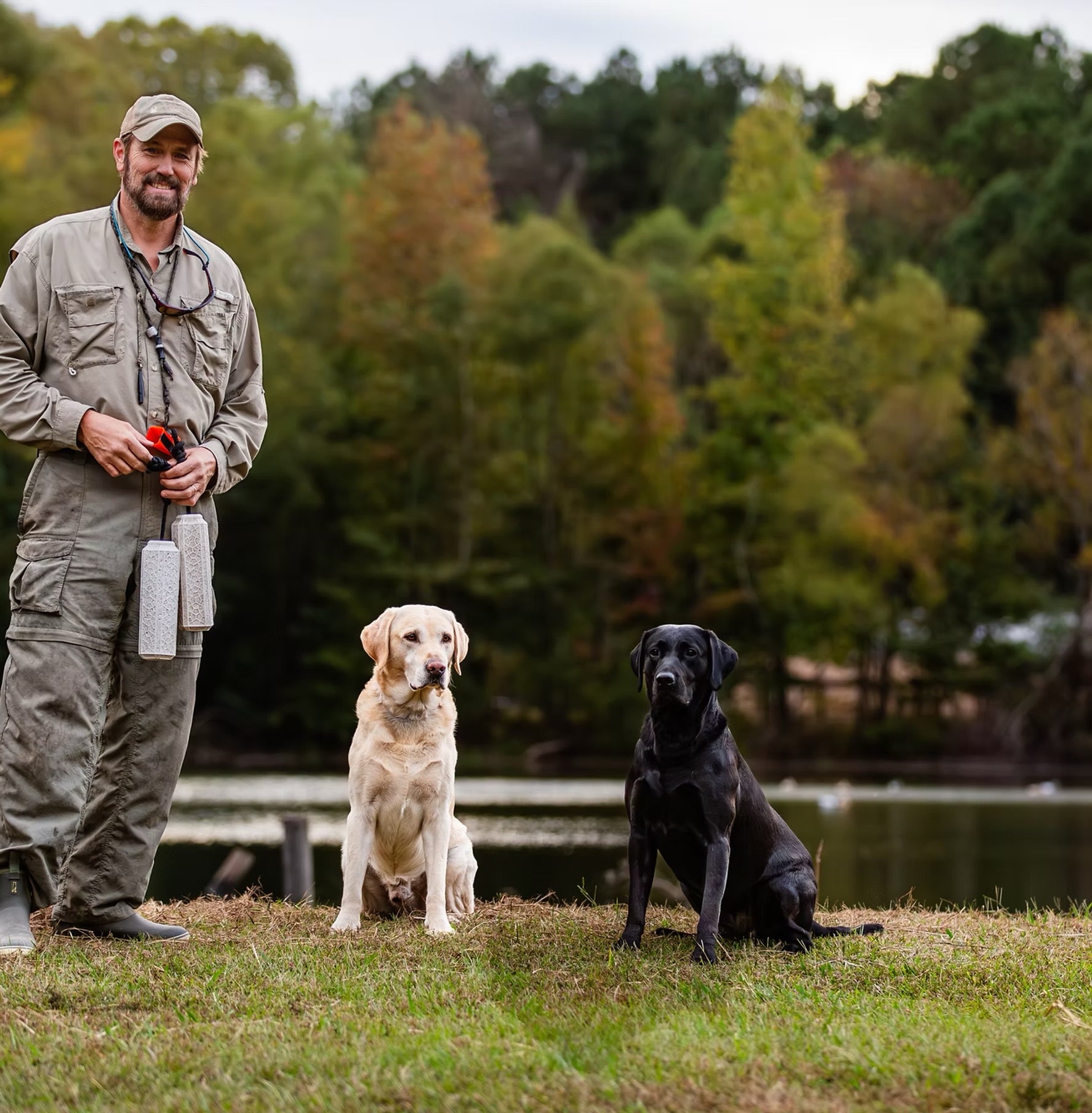 A handler with training dummies beside a yellow and a black Labrador, sitting at the edge of a pond.