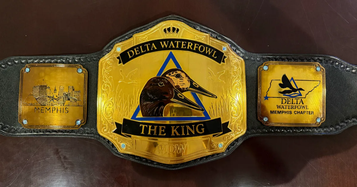 The finished championship belt on a dark wood table. A gold centre plate under a crown reads Delta Waterfowl above a blue triangle holding an engraved canvasback head, with a banner below reading The King. The left side plate is the Memphis skyline; the right carries the outline of Tennessee and the Delta Waterfowl Memphis Chapter mark.